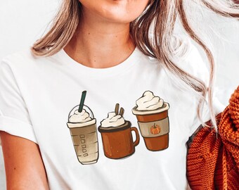 Puede incluir: Camiseta blanca con un gráfico de tres bebidas de café diferentes. Las bebidas son un latte de especias de calabaza, un caramel macchiato y un café frío. El gráfico está en estilo de dibujos animados.
