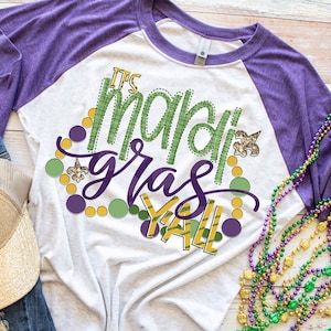 May include: A white and purple raglan t-shirt with the text "It's Mardi Gras Y'all" in green, purple, and gold. The shirt is decorated with green, purple, and gold beads and a gold fleur-de-lis.