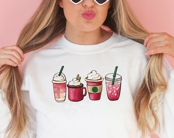 Puede incluir: Sudadera blanca con un gráfico de cuatro bebidas de café diferentes. Las bebidas están todas en vasos rojos con crema batida blanca y pajitas. Las bebidas incluyen un latte, un capuchino, un frappuccino y una bebida roja.