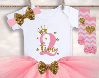 May include: Pink tutu with a white onesie featuring a gold crown and the number two with the word "two" underneath. The onesie also has gold glitter stars. The outfit includes a pink headband with a gold glitter bow and pink and white leg warmers with a gold glitter heart.