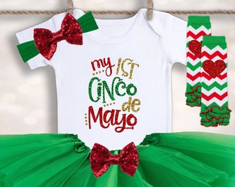May include: A green tulle tutu skirt, a white bodysuit with the text "my 1st Cinco de Mayo" in gold and red glitter, a red and green bow headband, and red and green leg warmers with a red heart design.