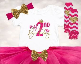 May include: A pink tulle skirt with a gold glitter bow, a white t-shirt with the text "my 2nd birthday" in pink and gold glitter, and pink and white striped leg warmers with a gold glitter heart.