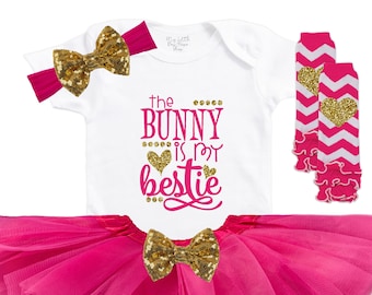 May include: A baby girl's outfit set featuring a white onesie with the text "the BUNNY is my bestie", a pink tutu skirt, a headband, and leg warmers. The set includes gold sequin bows and pink and white chevron leg warmers.