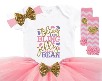 May include: A pink tulle tutu skirt with a white onesie that says "bling bling jelly bean" in purple and pink letters. The onesie has a gold glitter bow on the front. The outfit also includes a pink headband with a gold glitter bow and white and pink leg warmers with a gold glitter heart.