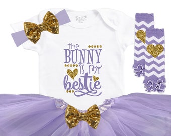 May include: A baby girl outfit set with a white bodysuit that says "The bunny is my bestie" in gold glitter lettering, a lavender tulle tutu, a lavender and white striped leg warmer with a gold glitter heart, and a lavender headband with a gold glitter bow.