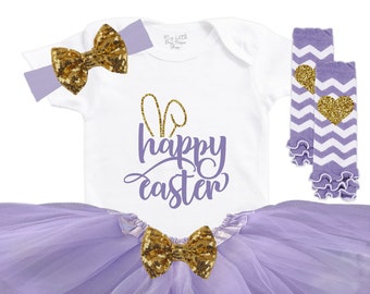May include: A baby girl's Easter outfit in purple and gold. The outfit includes a white bodysuit with bunny ears and the text "happy easter", a purple tulle skirt, a gold bow headband, and purple and white leg warmers with a gold heart.