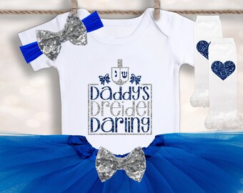Peut inclure: Une tenue de bébé fille pour Hanukkah. La tenue comprend un body blanc avec un motif pailleté argenté qui dit "Daddy's Dreidel Darling" et un tutu en tulle bleu. La tenue comprend également un bandeau à nœud pailleté argenté et des jambières blanches avec un motif de cœur bleu.