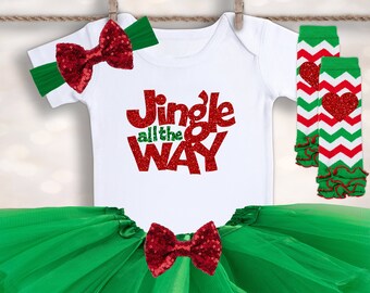 Puede incluir: Un tutú de tul verde con un lazo rojo, un body blanco con el texto "Jingle all the way" en purpurina roja y polainas a rayas rojas y blancas con un corazón rojo.