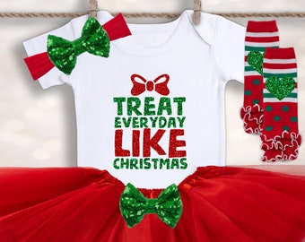 May include: A red tulle tutu skirt with a white bodysuit that says "Treat Everyday Like Christmas" in red glitter letters. The outfit is accessorized with a green glitter bow, a red headband with a green glitter bow, and red and green striped leg warmers with green glitter hearts.