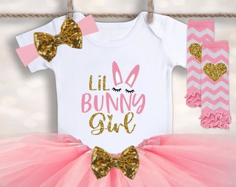 Puede incluir: Un tutú de tul rosa con un body blanco que lleva el texto "Lil Bunny Girl" y un conejito rosa con pestañas de purpurina dorada. El atuendo incluye una diadema rosa con un lazo de purpurina dorada y polainas rosa y blancas con corazones de purpurina dorada.