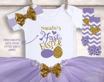 May include: A baby girl's first Easter outfit in shades of purple and gold. The outfit includes a white bodysuit with the text "Natalie's First Easter" and two gold glitter eggs, a purple tulle tutu, a purple headband with a gold glitter bow, and purple and white leg warmers with gold glitter hearts.