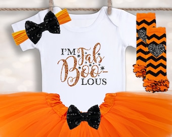 Może przedstawiać: Zestaw ubranek dla dziecka na Halloween, w tym białe body z napisem "I'm FabBOO-lous", pomarańczowa tutu, opaska na głowę i getry. Zestaw posiada czarne kokardy z cekinami oraz pomarańczowo-czarne wzory w szewrony.