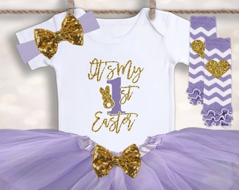 May include: A baby girl's first Easter outfit in lavender and gold. The outfit includes a white bodysuit with the text "It's My 1st Easter" in gold glitter, a lavender tulle tutu, a gold glitter bow headband, and lavender and white striped leg warmers with gold glitter hearts.