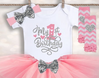 May include: A pink tulle tutu skirt with a silver sequin bow, a white onesie with the text "My 1st Birthday" in silver glitter lettering and pink hearts, a pink and white striped leg warmer with a silver heart, and a pink headband with a silver sequin bow.