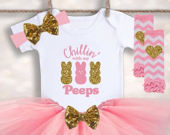 May include: A pink tulle tutu skirt with a gold glitter bow, a white bodysuit with the text "Chillin' with my Peeps" and three pink and gold glitter peep bunnies, and a pair of pink and white striped leg warmers with a gold glitter heart.