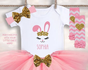 Puede incluir: Un conjunto para bebé niña con un body blanco con un diseño de cara de conejito rosa y el nombre "Sophia". El conjunto incluye un tutú rosa, un lazo de lentejuelas doradas, una diadema y calentadores de piernas con corazones dorados. El texto dice "¡PERSONALIZADO CON EL NOMBRE DE TU HIJO/A!"
