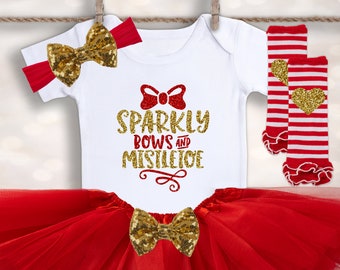 May include: A red tulle tutu skirt with a gold glitter bow, a white bodysuit with the text "Sparkly Bows and Mistletoe", and red and white striped leg warmers with gold glitter hearts.