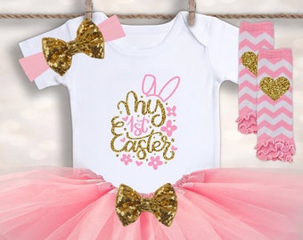 May include: A pink and gold Easter outfit for a baby. The outfit includes a white bodysuit with the text "My 1st Easter" in gold glitter, a pink tulle tutu, a pink headband with a gold glitter bow, and pink and white leg warmers with a gold glitter heart.