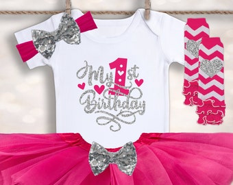 May include: A pink tutu, white onesie with the text "My 1st Birthday" in silver glitter, a pink and white chevron leg warmer, and a silver glitter bow headband.