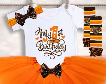 Op de afbeelding: Een witte romper met de tekst "My 1st Birthday" in oranje en bruin, gecombineerd met een oranje tutu, een haarband en beenwarmers. De haarband en tutu hebben een zwarte paillettenstrik. De beenwarmers hebben bruine en oranje strepen.