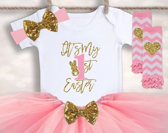 May include: Pink tulle tutu skirt with a gold glitter bow, a white onesie with the text "It's My 1st Easter" in gold glitter, and pink and white striped leg warmers with a gold glitter heart.