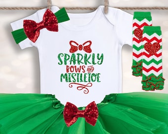 Puede incluir: Una falda de tutú de tul verde con un lazo rojo brillante, un body blanco con el texto "Sparkly Bows and Mistletoe" en purpurina roja, una diadema verde con un lazo rojo brillante y polainas a rayas rojas y blancas con un corazón rojo.