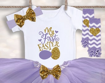 May include: A baby girl's Easter outfit in shades of purple and gold. The outfit includes a white bodysuit with the text "My First Easter" and a gold glitter egg, a purple tulle tutu, a gold glitter bow headband, and purple and white striped leg warmers with a gold glitter heart.