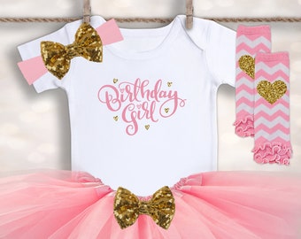 May include: Pink tulle tutu skirt with a gold glitter bow, a white bodysuit with the text "Birthday Girl" in pink glitter, and pink and white striped leg warmers with a gold glitter heart.