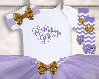 May include: A purple tulle tutu with a gold bow, a white bodysuit with the text "Birthday Girl" in purple script, a purple and white chevron leg warmer with a gold heart, and a gold bow headband.