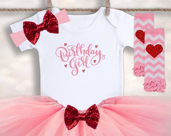 May include: A pink tulle tutu with a red sequin bow, a white onesie with the text "Birthday Girl" in pink script, and pink and white striped leg warmers with a red heart.