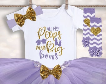May include: A baby girl's Easter outfit with a white bodysuit that says "All my peeps wear big bows", a purple tulle tutu, a gold bow headband, and purple and white chevron leg warmers with a gold heart.