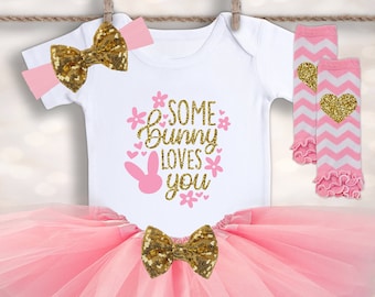 May include: A pink and gold baby girl outfit with a tutu skirt, a white bodysuit with the text "Some Bunny Loves You", a pink and gold headband, and pink and white leg warmers with gold hearts.