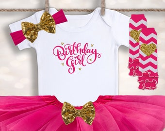 May include: A pink tulle tutu skirt with a white bodysuit that says "Birthday Girl" in gold glitter lettering. The outfit also includes a gold glitter bow headband and pink and white leg warmers with a gold glitter heart.