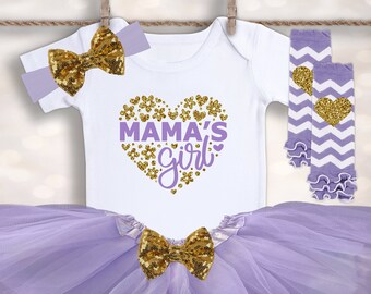 Könnte beinhalten: Ein Baby-Mädchen-Outfit-Set, einschließlich eines weißen Stramplers mit "MAMA'S girl" in einem Herz-Design, einem lavendelfarbenen Tutu-Rock, einem Stirnband und Beinwärmern. Das Set hat goldene Pailletten-Schleifen und Akzente.