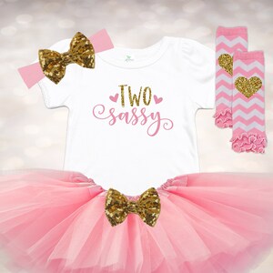 May include: Pink tulle tutu skirt with a gold glitter bow, a white t-shirt with the text "TWO sassy" in pink, a pink headband with a gold glitter bow, and white and pink leg warmers with a gold heart.