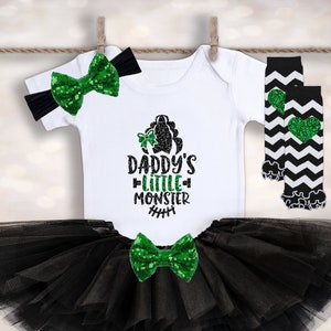 Op de afbeelding: Een zwarte tule tutu rok met een groene glitter strik. Een wit shirt met de tekst "Daddy's Little Monster" en een groene glitter strik. Zwart-wit gestreepte beenwarmers met een groen glitter hart.