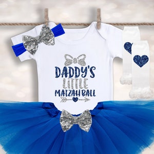 May include: A white bodysuit with the text "Daddy's Little Matzah Ball" in silver glitter. The bodysuit is paired with a blue tulle tutu, a silver glitter bow, and white leg warmers with a blue heart.
