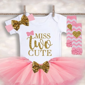 May include: A pink tutu with a gold glitter bow, a white onesie with the text "Miss Two Cute" in gold glitter, a pink headband with a gold glitter bow, and pink and white leg warmers with a gold glitter heart.