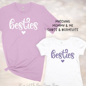 K&ouml;nnte beinhalten: Passende Mama-und-Ich-Shirts mit dem Text "besties" in Wei&szlig; auf einem lavendelfarbenen Shirt und in Lila auf einem wei&szlig;en Shirt. Beide Shirts haben ein kleines wei&szlig;es Herz.