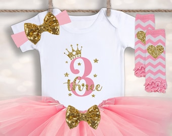 Può includere: Tutu rosa con un body bianco con una corona dorata e il numero 3 con la parola "three" sotto. Il body ha anche stelle dorate scintillanti. L'outfit include una fascia rosa con un fiocco dorato e gambaletti rosa e bianchi con un cuore dorato.