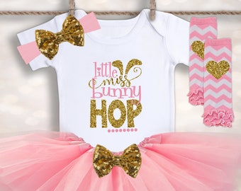 Puede incluir: Un conjunto de Pascua rosa y dorado para una niña pequeña. El conjunto incluye un body blanco con el texto "little miss bunny hop", un tutú de tul rosa, una diadema rosa con un lazo dorado y polainas rosa y blancas con un corazón dorado.