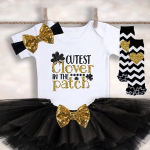 Könnte beinhalten: Ein schwarzes und goldenes Baby-Outfit mit einem weißen Body, auf dem "Cutest Clover in the Patch" steht. Das Outfit beinhaltet einen schwarzen Tüll-Tutu, ein goldenes Schleifen-Haarband und schwarze und weiße Beinstulpen mit goldenen Herzen.