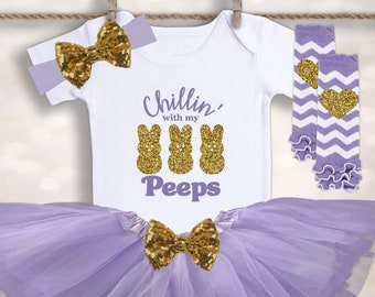 May include: A baby girl outfit with a white bodysuit that says "Chillin' with my Peeps" and has three gold glitter peeps on it. The outfit also includes a lavender tulle tutu with a gold glitter bow, a lavender headband with a gold glitter bow, and lavender and white leg warmers with a gold glitter heart.