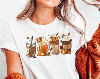 Puede incluir: Camiseta blanca con un gráfico de cuatro bebidas con temática otoñal. Las bebidas son un latte de especias de calabaza, una cerveza de calabaza, una sidra de manzana con caramelo y un latte de pastel de calabaza. Cada bebida está cubierta con crema batida y una calabaza. El gráfico está rodeado de hojas de otoño y palitos de canela.