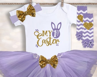 May include: A baby girl's Easter outfit in shades of purple and gold. The outfit includes a white bodysuit with the text "My 1st Easter" and a bunny rabbit graphic, a purple tulle tutu, a purple and white chevron leg warmer with a gold heart, and a purple headband with a gold bow.