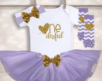 Op de afbeelding: Een baby-outfit voor de eerste verjaardag in wit, lavendel en goud. De set bevat een body met gouden glittertekst, een tutu rok, een haarband en beenwarmers. De tekst op de body luidt "One derful". De beenwarmers hebben een chevron patroon.