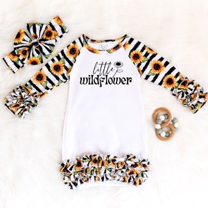 Pode incluir: Batista branca para bebê com mangas compridas e acabamento de babado. As mangas e o babado inferior são listrados em preto e branco com estampa de girassol. A bata tem o texto "little wildflower" impresso na frente.