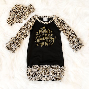 Puede incluir: Batista de bebé negra con ribete de estampado de leopardo y mangas largas. La bata tiene un diseño de purpurina dorada que dice "Brand Sparkling New" con un corazón. Se incluye una diadema a juego con estampado de leopardo.