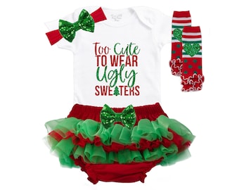 Puede incluir: Un body blanco con el texto "Too Cute To Wear Ugly Sweaters" en purpurina roja. El body se combina con un tutú de tul rojo y verde, un lazo de purpurina verde y polainas a rayas rojas y verdes con un corazón de purpurina verde.