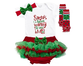 Puede incluir: Un body blanco con el texto "Santa's been waiting to see ME!" en purpurina roja y verde. El body se combina con un tutú de tul rojo y verde, un lazo de purpurina verde y polainas a rayas rojas y verdes con un diseño de corazón.